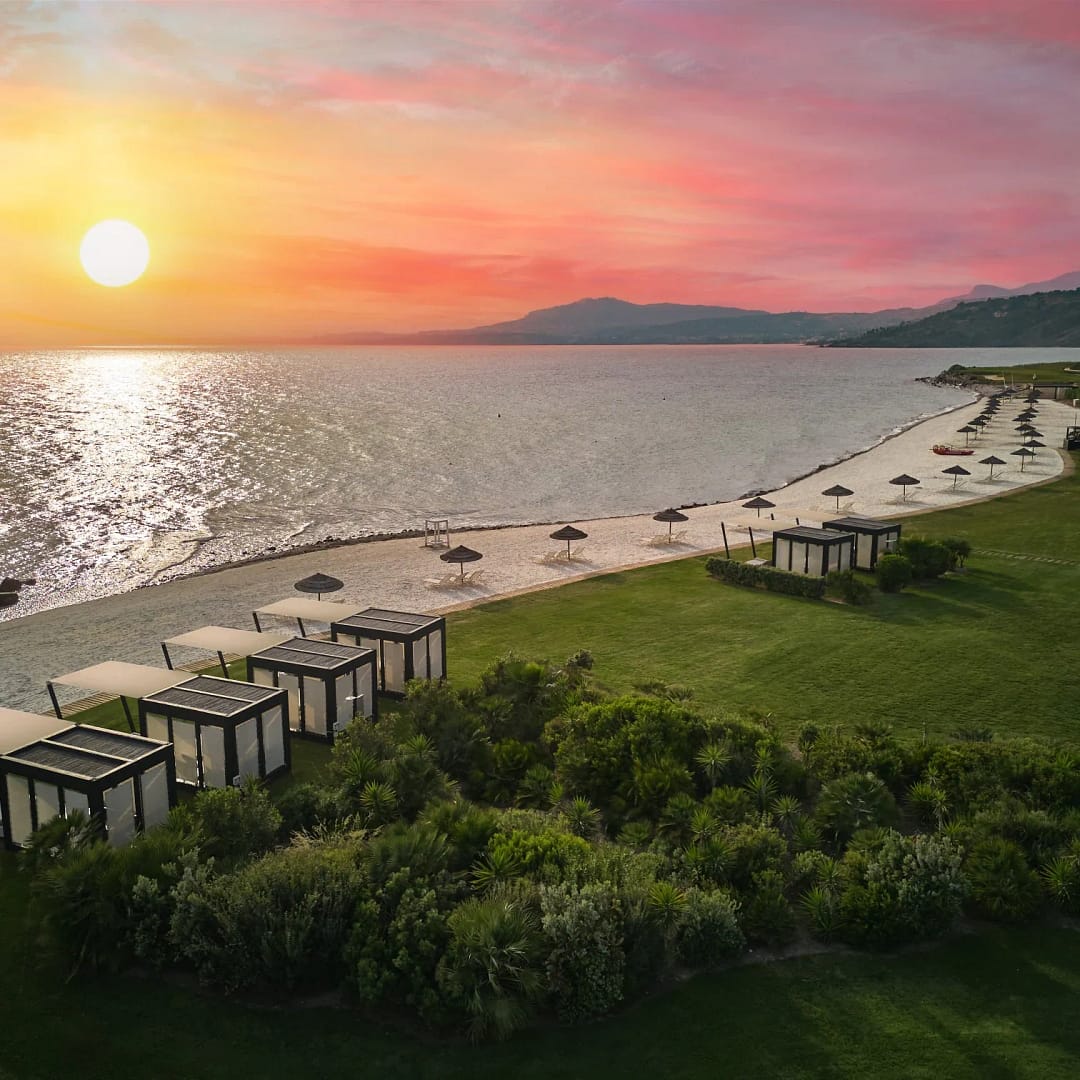 Long pale beach with umbrellas.  Lawn behind the beach. Sea to the left with sunset and red sky.