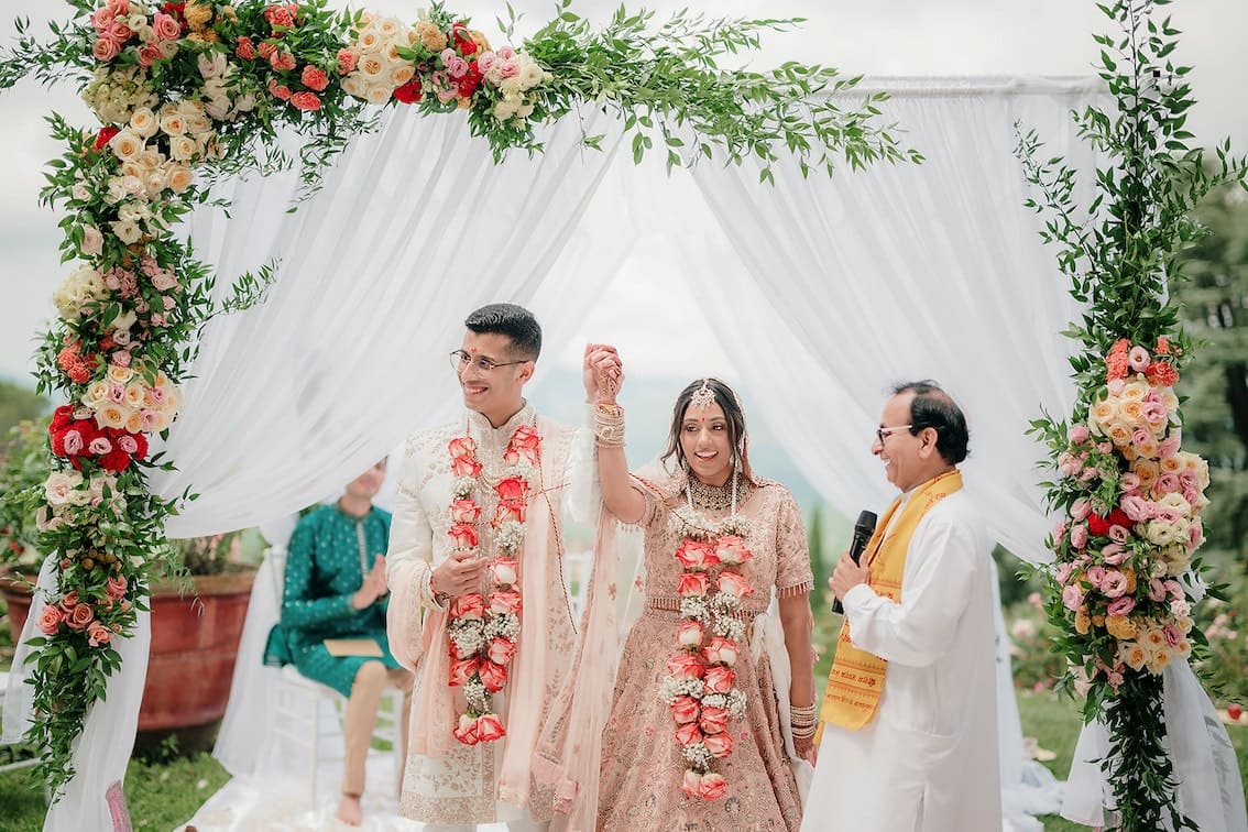 Indian couple under a mandap celebrating after their wedding ceremony. They have flowers around their shoulders and look very happy.