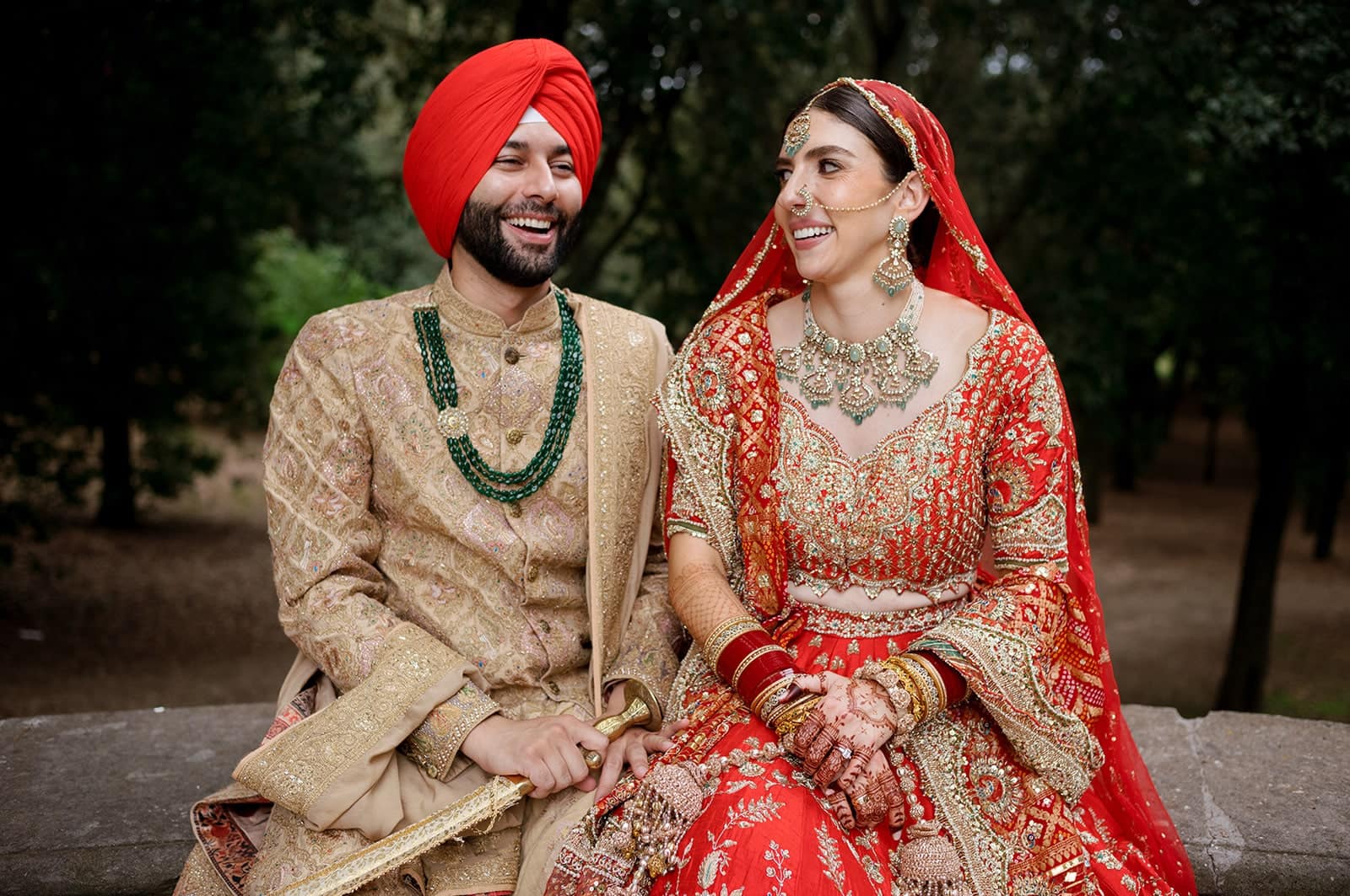 Indian bride and groom pose for a photo in their wedding attire. She has a red sari, he a red turban and a gold jacket. They are sitting down.