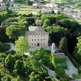 Square shaped Italian villa made from pale brick, surrounded by green trees.