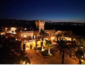 Masseria in Puglia photographed at night.  It is illuminated by yellow lighting.  There are palm trees in the foreground and an inky blue night sky in the background.