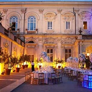 Italian palace facade with dining tables set up for a wedding in front.  It is dusk and the scene is illuminated by coloured lights.