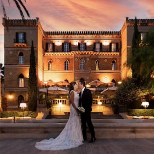Bride % groom stand in front of an elegant palace in Palermo.  It is sunset and they have their wedding at the palace.