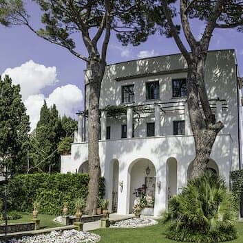 Front of a white villa with an arched portico.  The villa has three storeys.  There is a lawn in front with a path leading to the portico.
