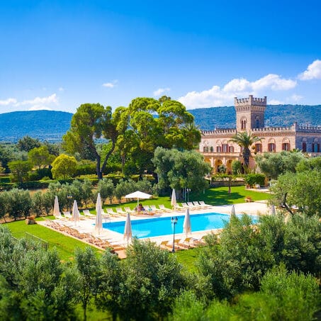 Masseria in Puglia with castelated roof and a tower.  It is set in green gardens with very large green shrubs and a few trees.  There is swimming pool.