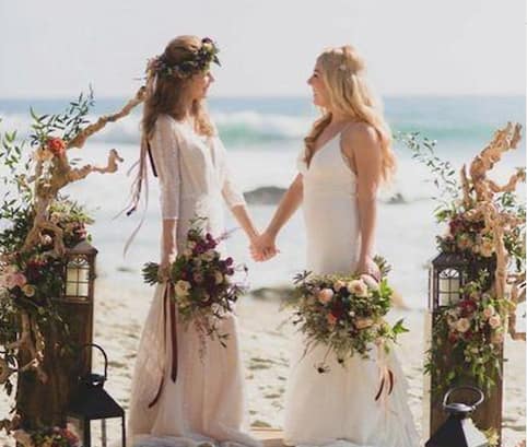 Two women hold hands and look into each other's eyes as they marry in a gay beach wedding. The sea is behind them and they are standing on the sand. There are floral compositions to each side of them creating a wedding alter type space.