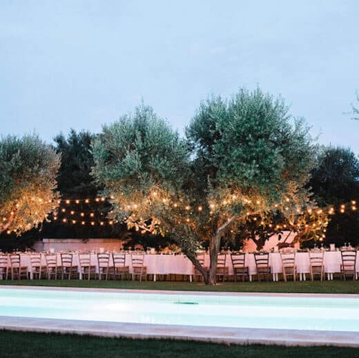 long dining table set up for a wedding.  In an olive growth with fairy lights in the olive trees.  Swimming pool in foreground.