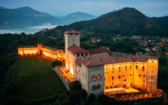 Aerial view of castle wedding venue, Rocca di Angera in Italy. It is pictured in twilight. The castle is in the foreground and the facade is illuminated with a warm orange light. In the background you can see Lake Maggiore.