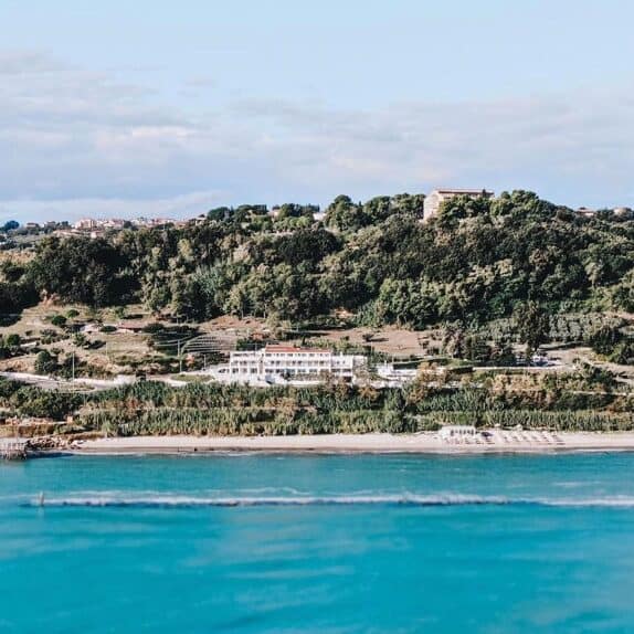 A green hillside leading down to the sea.  The sea is in the foreground.  There's a large white building at the bottom of the hill.