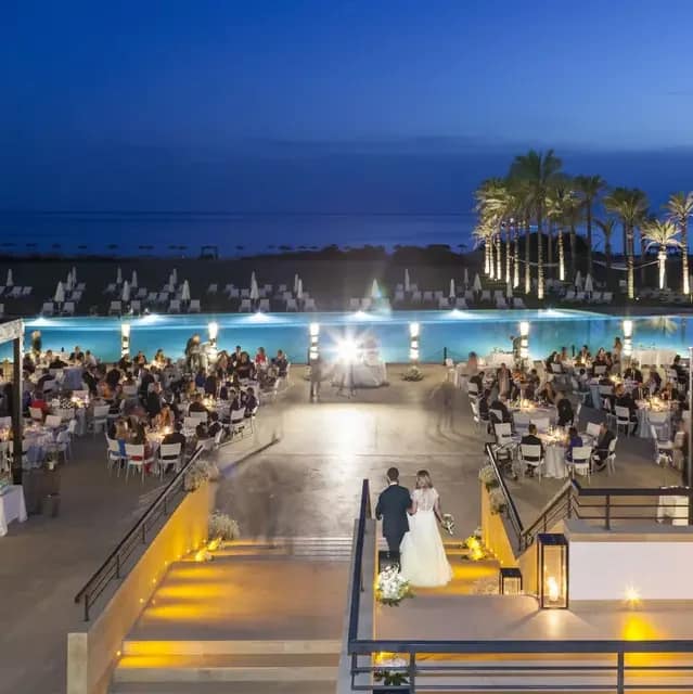 A luxurious swimming pool with a large terrace.  Dining tables set up for a wedding banquet.  It is dusk.  The illuminated swimming pool  is behind, with the beach behind that.