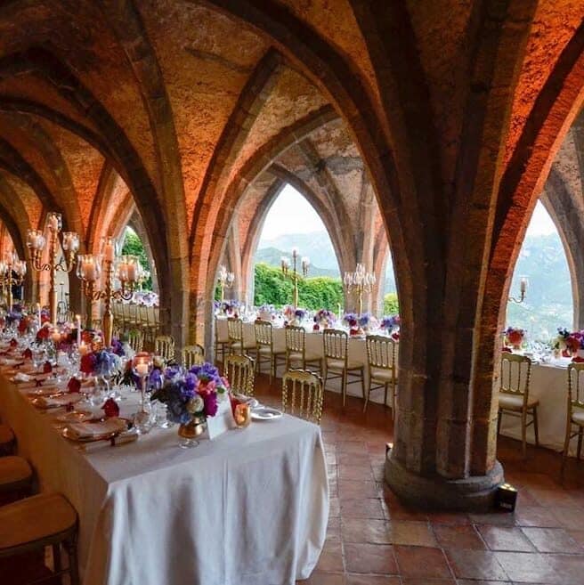 Antique vaulted hall set up with dining tables prepared for a wedding banquet.  The far wall is open and there is a view of the Amalfi coast.