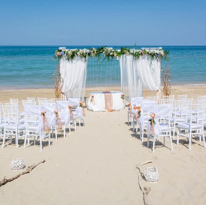 Wedding ceremony on a beach.  There is a white decorated arch in front of rows of white chairs.