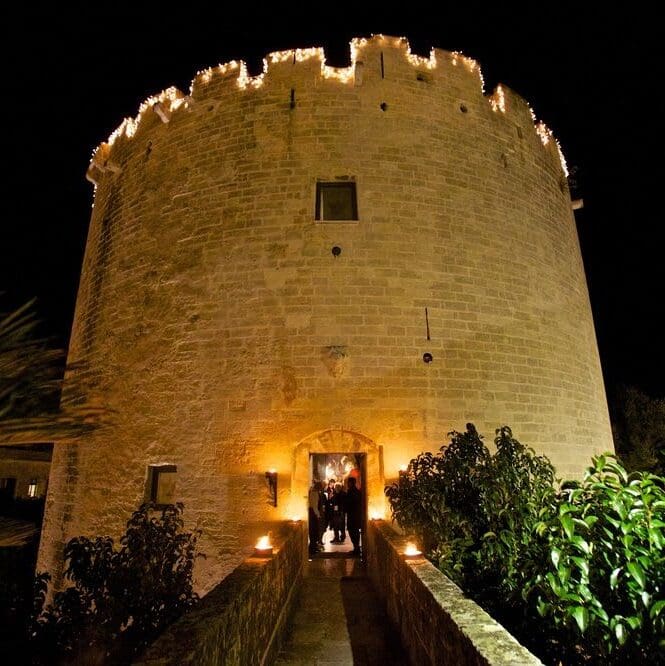 A medieval tower photographed from below at night.  There are yellow lights around the door and on the castellated roof.