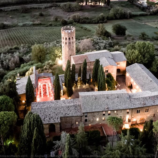 Aerial view of historic Italian abbey.  There are stone buildings around a central courtyard.  Tall conifers growing out of the courtyard.  There is a very tall tower.