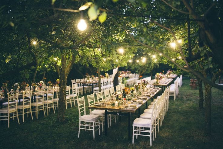 Wedding dining tables set up under trees. It is night time and there is yellow light from overhead fairy lights.