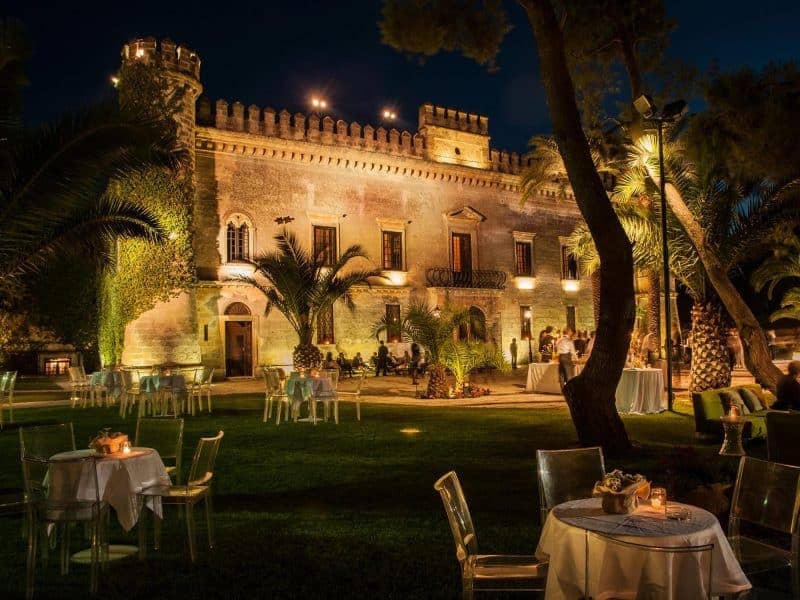 Facade of an Italian castle with palm trees in foreground.