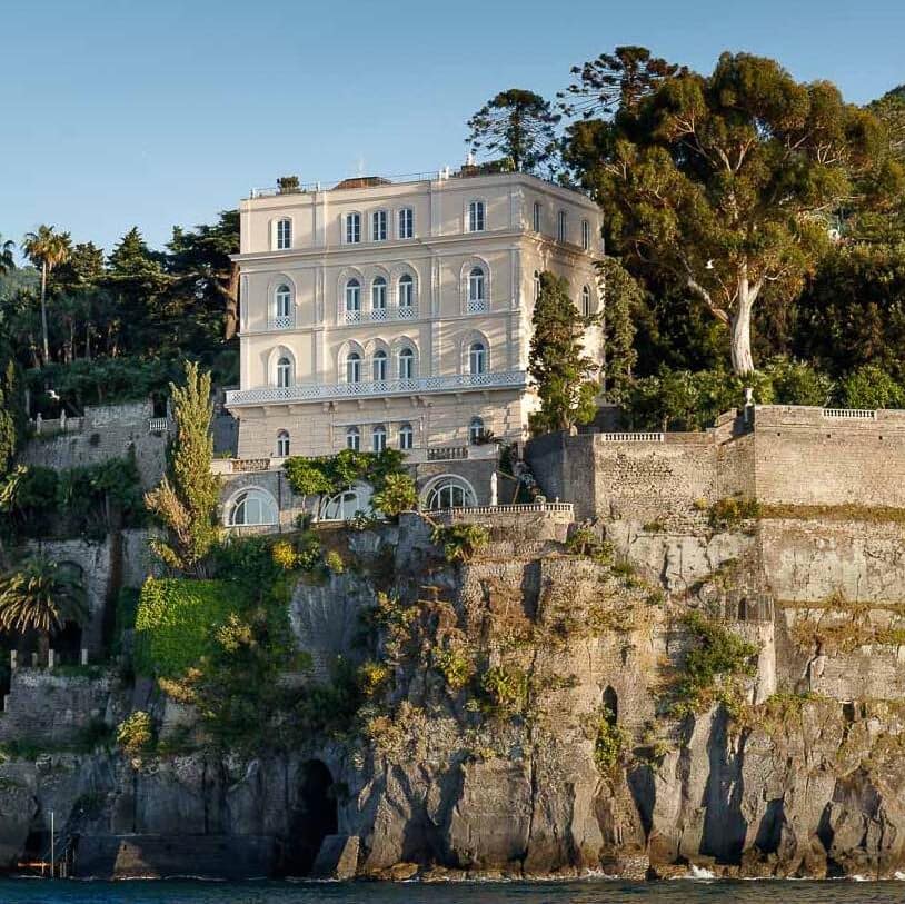 Tall cream coloured villa on top of a cliff above the sea.