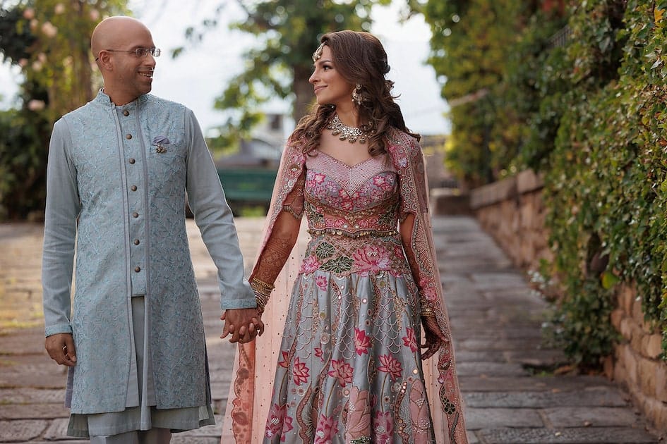 Happy Indian couple walk hand in hand and smiling. They wear blue & pink traditional Indian outfits.