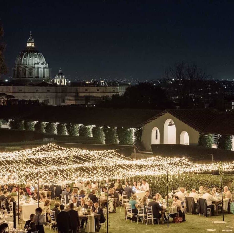Villa Piccolomini. Wedding venue in Rome, Italy. View of large lawn set up with dining tables under fairy lights. View of St Peter's in the background.